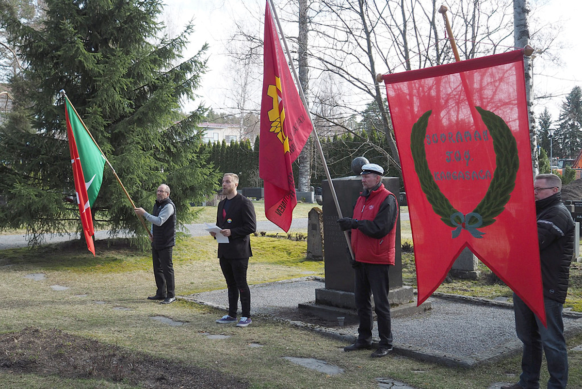 Vapputilaisuus Kangasalan punaisten muistomerkillä. Kolme miestä pitelee työväen lippuja ja yksi pitää puhetta.