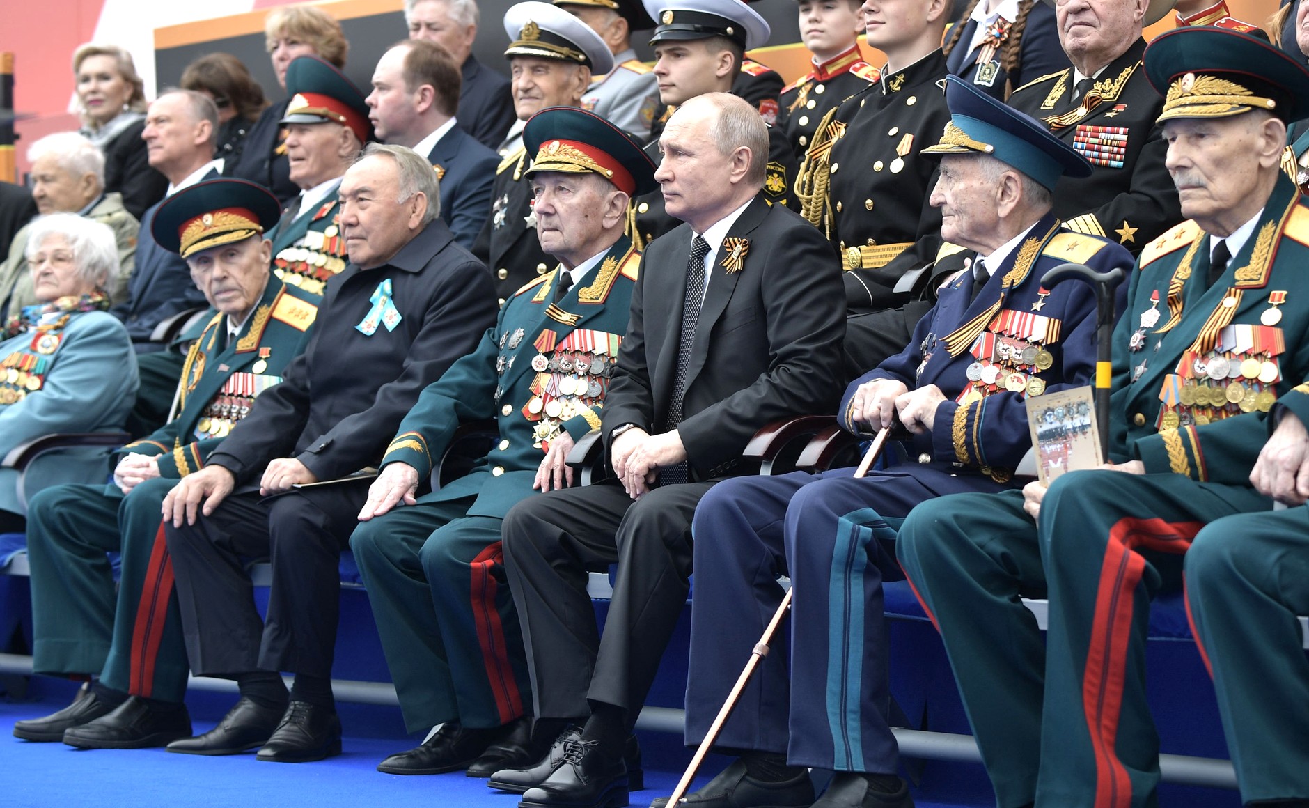 Venäjän presidentti Vladimir Putin istuu sotaveteraanien ja Kazakstanin presidentin kanssa katsomossa Voiton päivän paraatissa vuonna 2019.