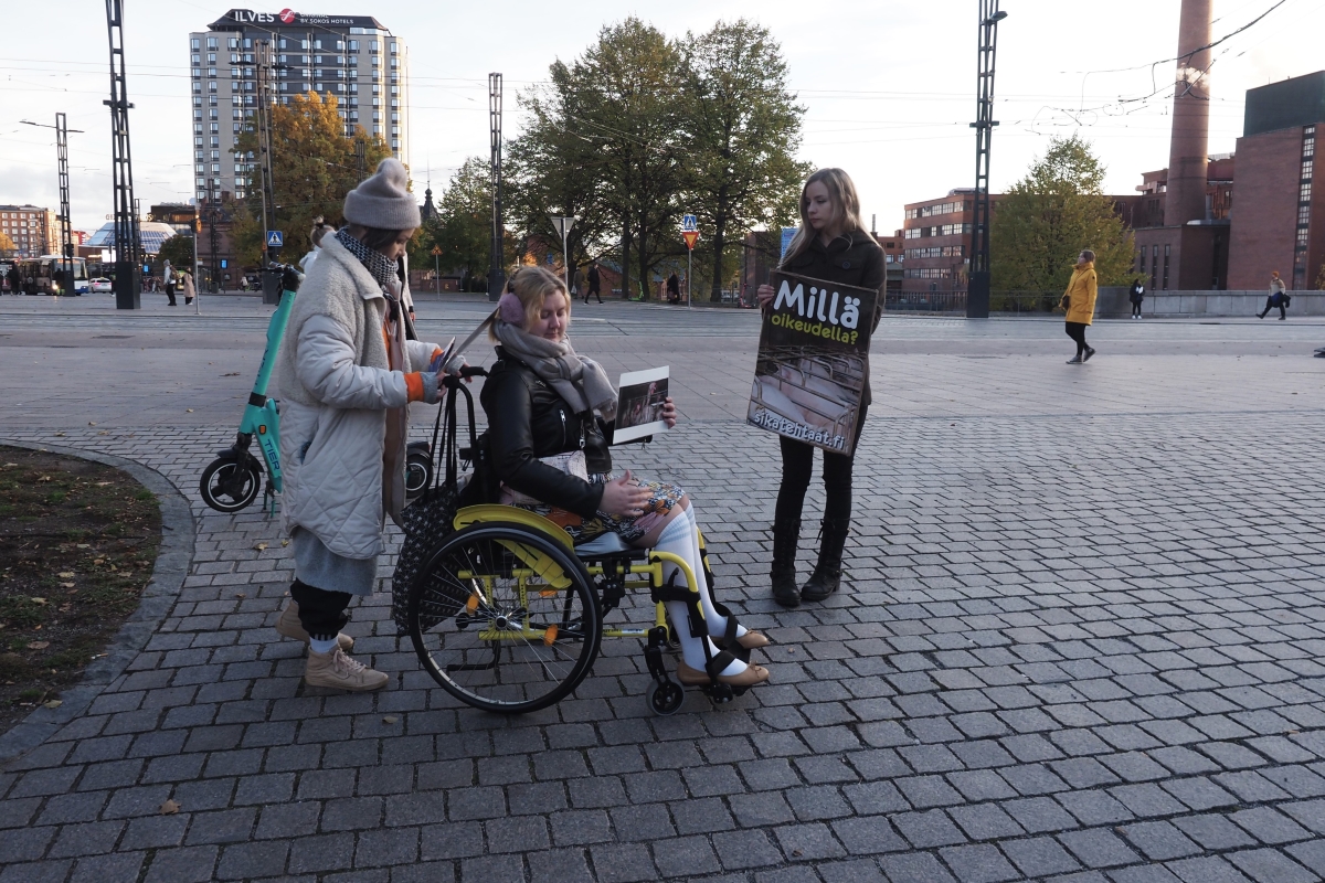 Kolme henkilöä aukealla kadun vieressä. Keskellä oleva istuu pyörätuolissa, muut seisovat.