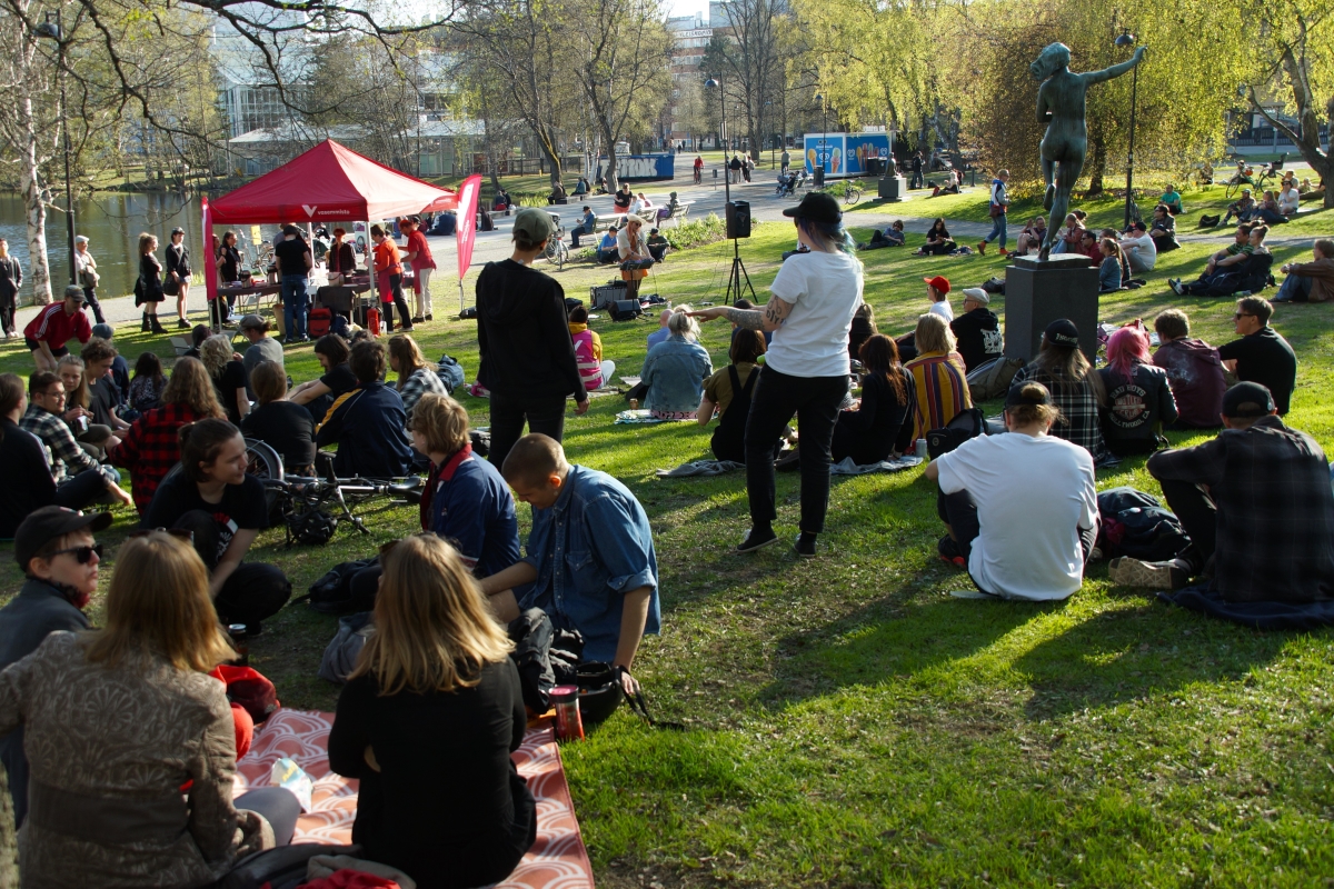 Ihmisiä istumassa nurmikolla Sorsapuistossa, edessä vasemmiston teltta ja laulaja.