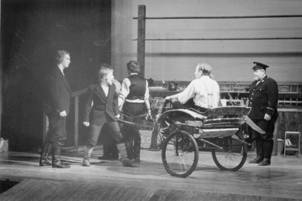 Mustavalkoinen kuva näyttämöltä, jossa yksi näyttelijä istuu kieseissä, yksi seisoo vieressä ja kaksi edessä. Tilanne näyttää alkavalta tappelulta. 