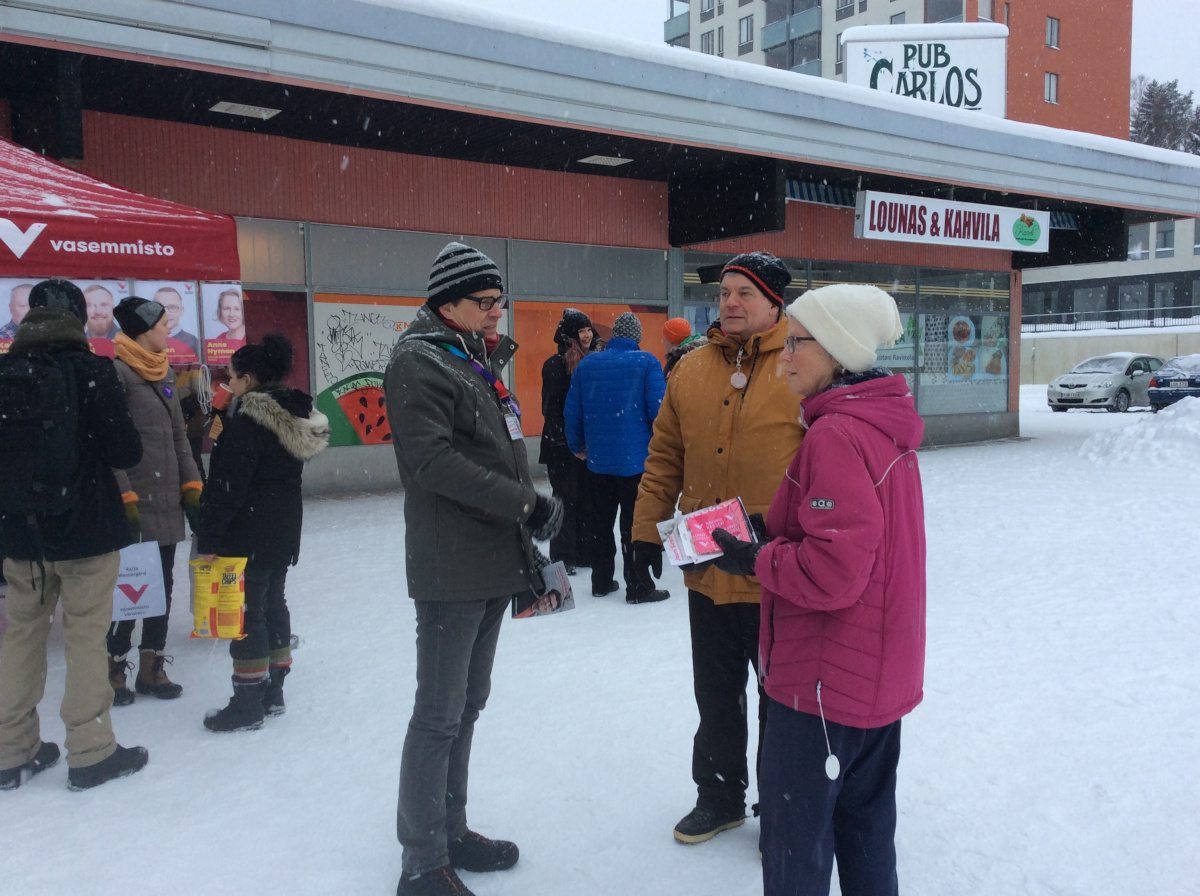 Kolme ihmistä keskustelee talvisessa maisemassa. Taustalla ostoskeskus.